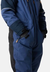 Blå isolerad overall med svarta sidopaneler, utrustad med dragkedjefickor, förstärkta sömmar och reflekterande detaljer på axlarna.