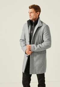 Cappotto grigio in lana con motivo a spina di pesce, dotato di colletto alto, bottoni frontali e tasche laterali, abbinato a uno strato interno nero.