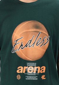 Grøn bomulds T-shirt med et stort orange basketballgrafik og teksten "Endless" ovenfor, samt orange og hvid tekst forneden.