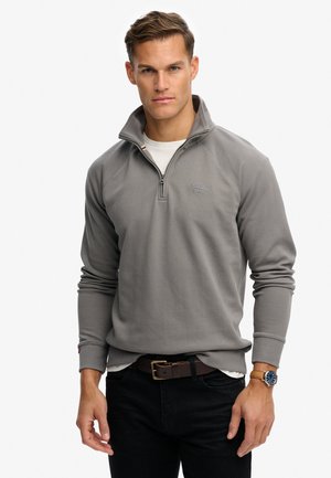 Sweat à zip gris avec un col haut et des poignets côtelés, arborant un petit logo sur la poitrine, associé à un jean noir et une ceinture marron.