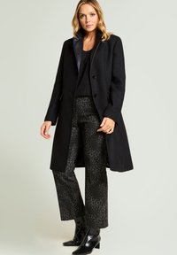 Abrigo de lana negra con cuello negro brillante, combinado con pantalones negros con estampado. El abrigo presenta cierres de botones y un ajuste entallado.