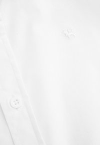 Biała koszula wykonana z gładkiej tkaniny, z małym haftowanym logo. Na kołnierzyku widoczny jest jeden biały guzik.