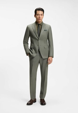 Mężczyzna stojący z rękami w kieszeniach, ubrany w jasnoszary garnitur, oliwkowo-zieloną koszulę, czarny krawat i ciemnobrązowe eleganckie buty na białym tle.