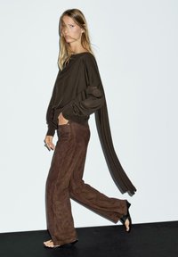 Brown knitted long-sleeve top with draped design, paired with brown suede wide-leg pants. Model wearing black sandals, standing on a dark surface.