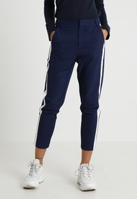 Marinblå byxor med vita sidränder, åtsittande design, slät textur och kort längd, bärs med vita sportskor.