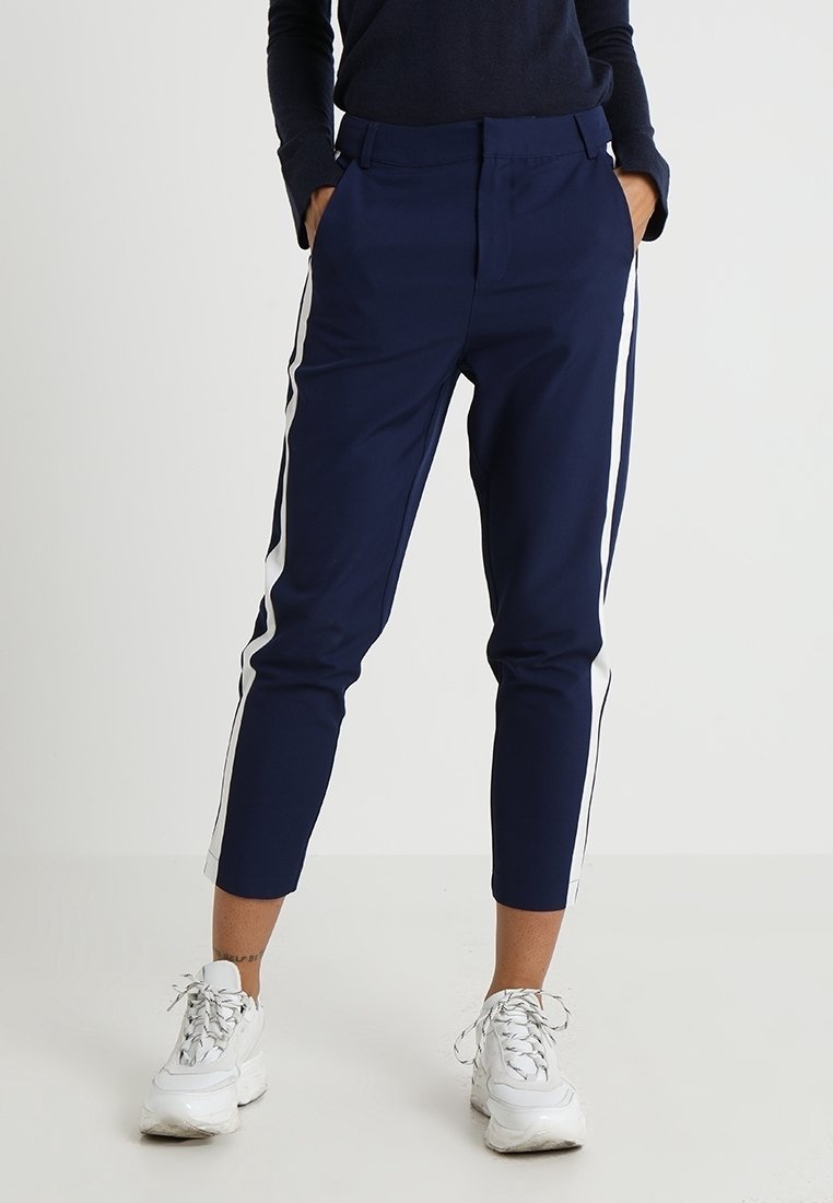 Marinblå byxor med vita sidränder, åtsittande design, slät textur och kort längd, bärs med vita sportskor.