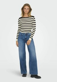 Randig långärmad topp i svart och krämbelägg, kombinerad med högmidjade blå vida jeans. Outfitten har ribbad textur och stiliga svarta skor.