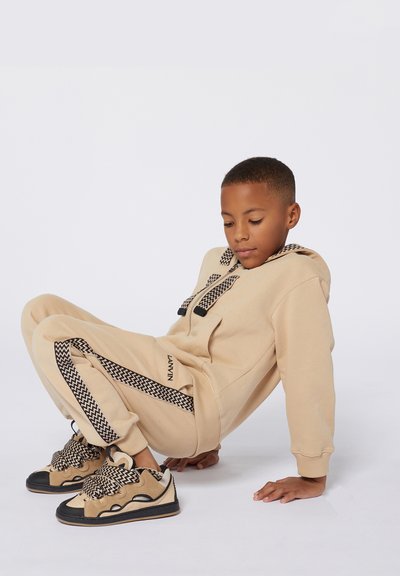 Sweatshirt beige avec capuche en chevrons, pantalons assortis avec des rayures sur les côtés. Les baskets présentent un design superposé en beige et noir.