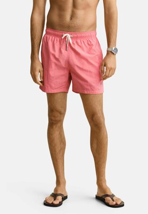 Man draagt koraalroze zwembroek met witte trekkoord, een zilveren horloge en donkergroene slippers, staand tegen een effen witte achtergrond.