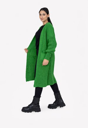 Vrouw die loopt en een lange groene gebreide cardigan draagt over zwarte kleding en zwarte veterlaarzen, tegen een effen witte achtergrond.