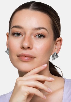 Kobieta z zaczesanymi do tyłu włosami, nosząca marmurkowe srebrne trójkątne kolczyki, opierająca podbródek na ręku, ubrana w jasnofioletowy top.