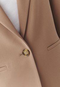 Blazer marrone realizzato in tessuto liscio, con un grande bottone rotondo beige, reverse a lancia e una tasca sul petto.