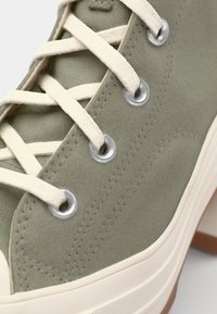 Zapatilla alta de lona verde con cordones crema, ojales plateados y una superficie de tela texturizada. Presenta una suela de goma y detalles de costura.