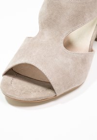 Sandal in suede beige con tacco alto, caratterizzata da punta aperta, design con ritagli e interno liscio. La soletta è di colore chiaro con cuciture visibili.