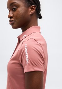 Γυναίκα με ροζ πουκάμισο με γιακά και κοντά μανίκια, με την επιγραφή "J.LINDEBERG" σε λευκό χρώμα στο αριστερό μανίκι, στραμμένη προς τα αριστερά σε απλό φόντο.