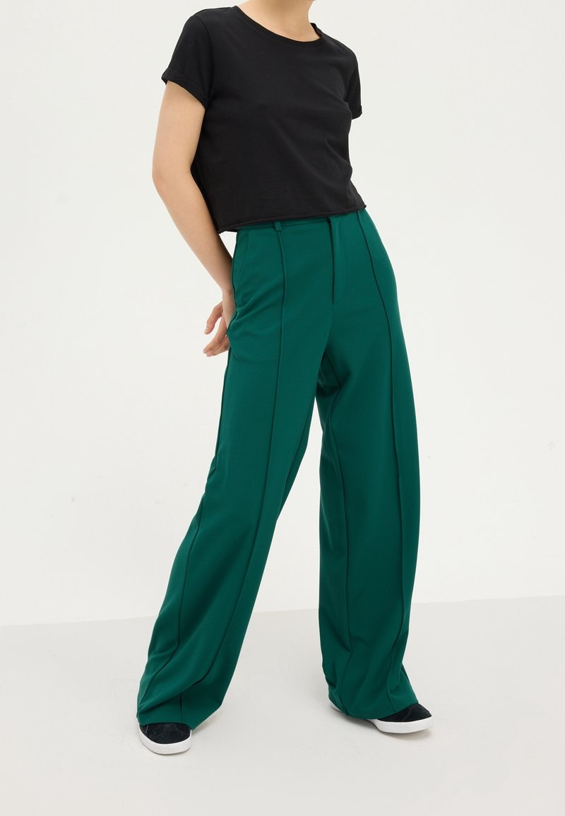 Personne portant un pantalon vert à jambes larges, un t-shirt noir court et des chaussures noires à enfiler, debout sur un fond blanc.