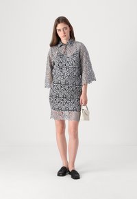 Lacekjole i svart og sølv med korte ermer, krage-design og scallop-fall. Modellen holder en liten hvit håndveske. Svart loafers er brukt.