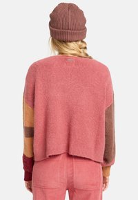 Fluffy roze trui met meerkleurige patroonmouwen, gecombineerd met een bruine geribbelde beanie. Zachte textuur, cropped ontwerp en relaxte pasvorm.