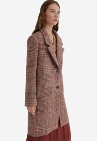 Kvinde iført en lang, knaplukket ternet frakke i lyserøde, brune og orange nuancer over en bordeaux nederdel, med lagdelte guldhalskæder.