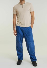 Beige polo triko s krátkým rukávem a decentním logem; modré cargo kalhoty s velkými našitými kapsami a roztrhanými okraji; černé tenisky.