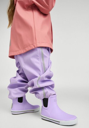 Dziecko ubrane w różową kurtkę, fioletowe wodoodporne spodnie z odblaskowymi paskami oraz pasujące fioletowe kalosze, stojące na szarym tle.