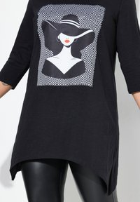 Tunic nera con una stampa grafica di una donna che indossa un cappello a tesa larga. Maniche a tre quarti e orlo asimmetrico. Tessuto leggero.