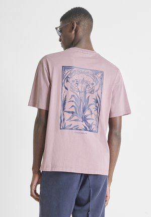 Muškarac odjeven u svijetloružičastu majicu s plavim botaničkim uzorkom i natpisom „MEZCAL“ na leđima, uparen s tamnoplavim hlačama, okrenut prema suprotnoj strani.