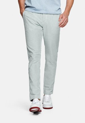 Homme portant un pantalon slim à fines rayures bleu clair et blanc, avec des baskets blanches à semelles rouges, marchant vers l'avant sur un fond blanc.