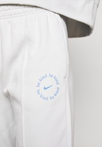 Ljusgrå svettbyxor i mjukt material, med en blå cirkulär logotyp som har upprepad text "var snäll" och en Nike-swoosh.