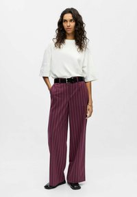 T-shirt blanc surdimensionné à manches courtes, associé à un pantalon large bordeaux à rayures et une ceinture noire. Des chaussures noires viennent compléter la tenue.