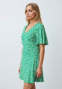Groene jurk met witte bloemenprint, V-hals en fladdermouwen. Gemaakt van lichtgewicht stof, heeft het een uitlopende silhouet.