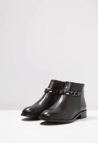 Botas de tobillo de cuero negro con punta redonda, tacones planos, cremalleras laterales y una cadena decorativa de metal negro en la parte delantera.