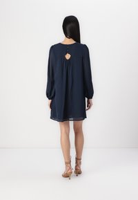 Vestido azul-marinho, até aos joelhos, com mangas longas, detalhe em forma de buraco na parte de trás e três botões no decote. Tecido leve e texturizado.