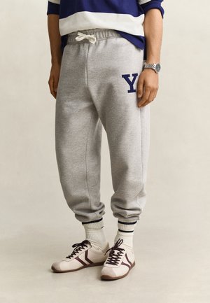 Persona que lleva pantalones deportivos grises con un logo "Y" azul marino, calcetines blancos altos con rayas azul marino, zapatillas blancas con detalles marrones y un reloj de pulsera plateado.