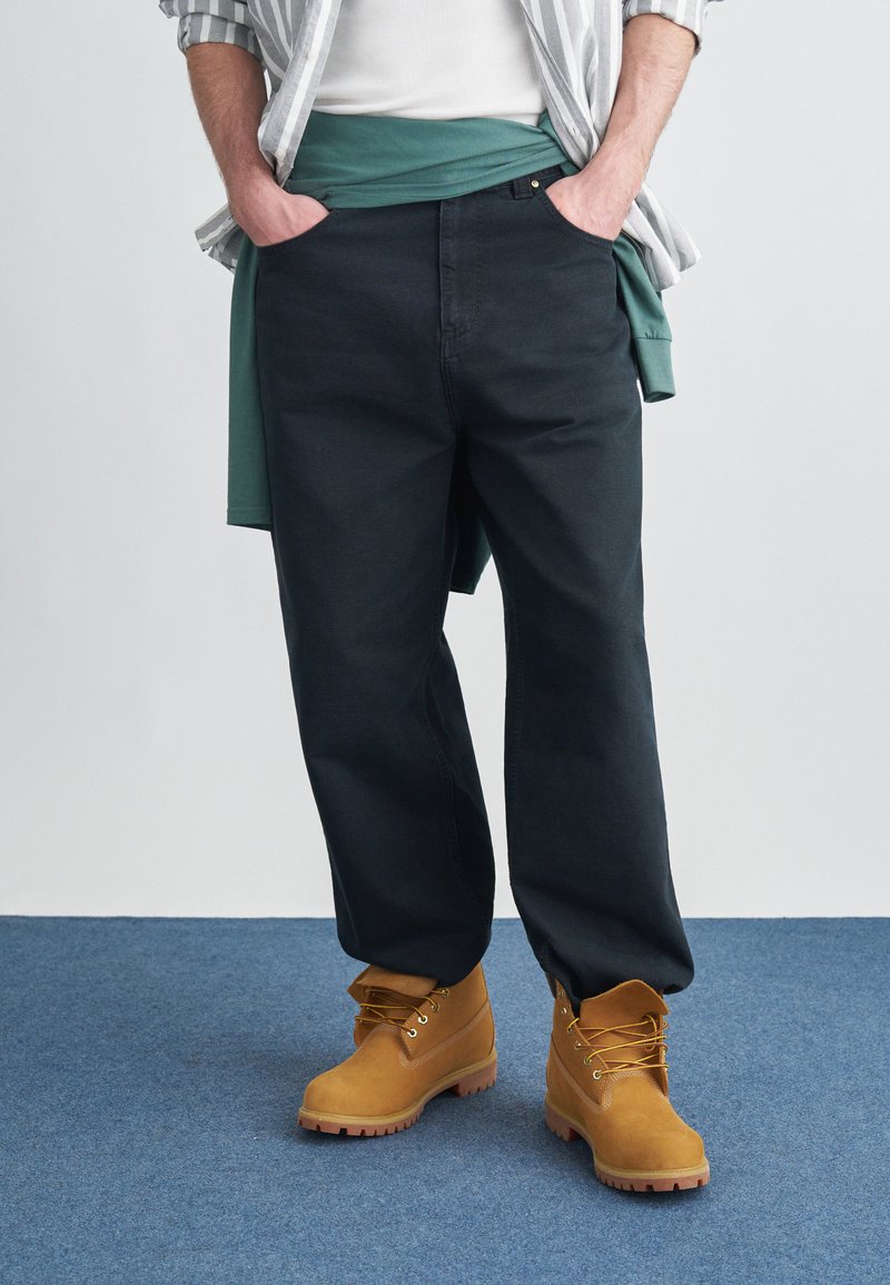 Σκούρο μπλε παντελόνι σε χαλαρή εφαρμογή, συνδυασμένο με πράσινο φούτερ δεμένο στη μέση και κίτρινο-καφέ μποτάκια με κορδόνια και υφή στη σόλα.