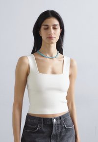 Λευκό, εφαρμοστό αμάνικο μπλουζάκι με τετράγωνη λαιμόκοψη, συνδυασμένο με ψηλόμεσα σκούρα τζιν. Απλός σχεδιασμός, ομαλή υφή, χωρίς ορατά μοτίβα.