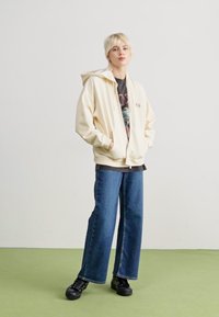 Kremfarget zip-up hettegenser med avslappet passform, med stor hette og frontlommer. Bært over en mørk grafisk t-skjorte og vide jeans.