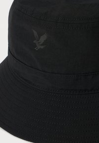 Schwarzer Bucket Hat mit genähtem Rand und einem kleinen schwarzen Adler-Logo an der Seite.