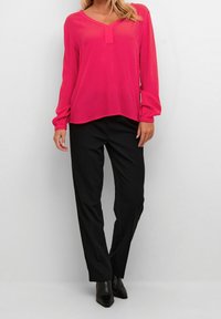 Blusa rosa a maniche lunghe con scollo a V e dettagli con bottoni, abbinata a pantaloni neri a sigaretta e stivaletti neri a punta. Tessuto dalla texture liscia.