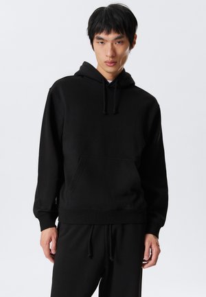 Young man wearing black hoodie and black sweatpants standing against white background, looking forward with relaxed posture.