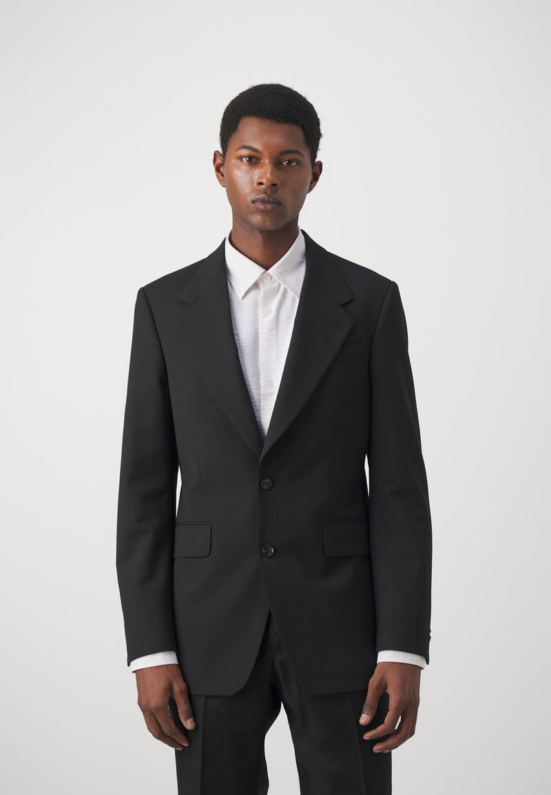 Zwarte op maat gemaakte blazer met gelapte revers, twee knopen en ingeweven zakken. Draagt over een witte overhemd met kraag en presenteert een gestructureerd silhouet.