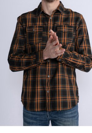 Mann trägt ein schwarz-orange kariertes Hemd mit Knöpfen und blaue Jeans, hält die Hände vor der Brust verschränkt vor einfachem Hintergrund.