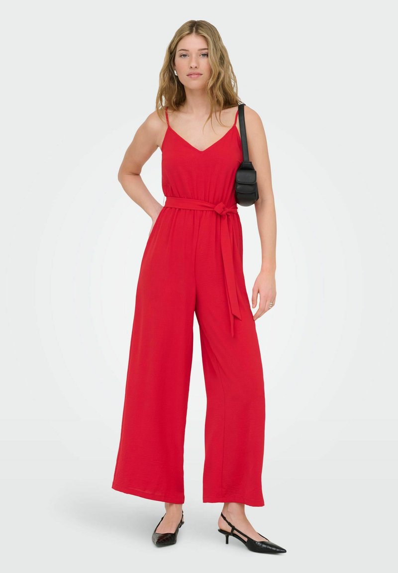 Tânără purtând un combinezon roșu fără mâneci, cu picioare largi, pantofi negri cu barete în spate și o geantă mică neagră de umăr.