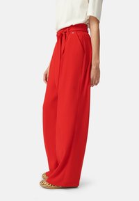 Vrouw in hoogwaist wijd-uitlopende rode broek met een gestrikt ceintuur, witte top en luipaardprint instappers, staande in zijaanzicht.
