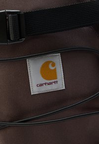 Καφέ ύφασμα με ετικέτα λογότυπου Carhartt, μαύρο ρυθμιζόμενο λουράκι και μαύρα κορδόνια σύσφιξης σε τσάντα ή σακίδιο.