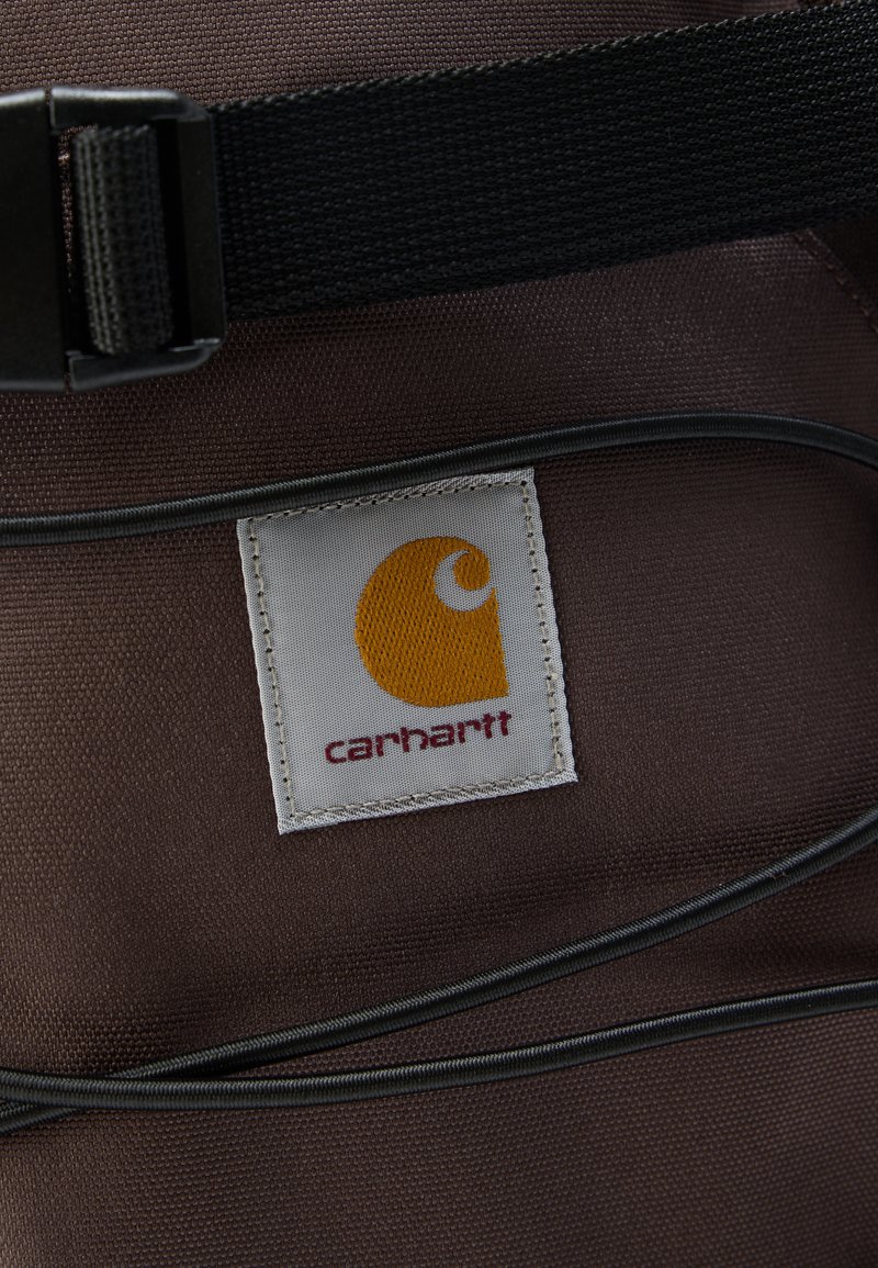 Bruin stof met Carhartt-logo patch, zwarte verstelbare riem en zwarte trekkoorden aan een tas of rugzak.