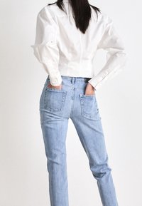 Person med långt mörkt hår bär en vit skjorta och ljusblå jeans, med händerna stoppad i bakfickorna, stående mot en enkel vit bakgrund.