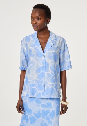 Vrouw draagt een lichtblauw tie-dye kort mouwen blouse met knopen en bijpassende rok, gecombineerd met een crème armband.