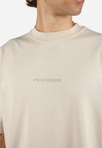 Beige bomull T-skjorte med rund hals og korte ermer. Har en subtil "PEGADOR"-logo i en mørkere nyanse på brystet. Jevn tekstur.