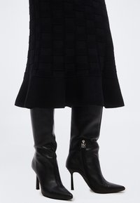 Μαύρες δερμάτινες μπότες μέχρι το γόνατο με μυτερά δάχτυλα και τακούνια στιλέτο, συνδυασμένες με μαύρη υφασμάτινη φούστα που τελειώνει στη μέση της γάμπας.